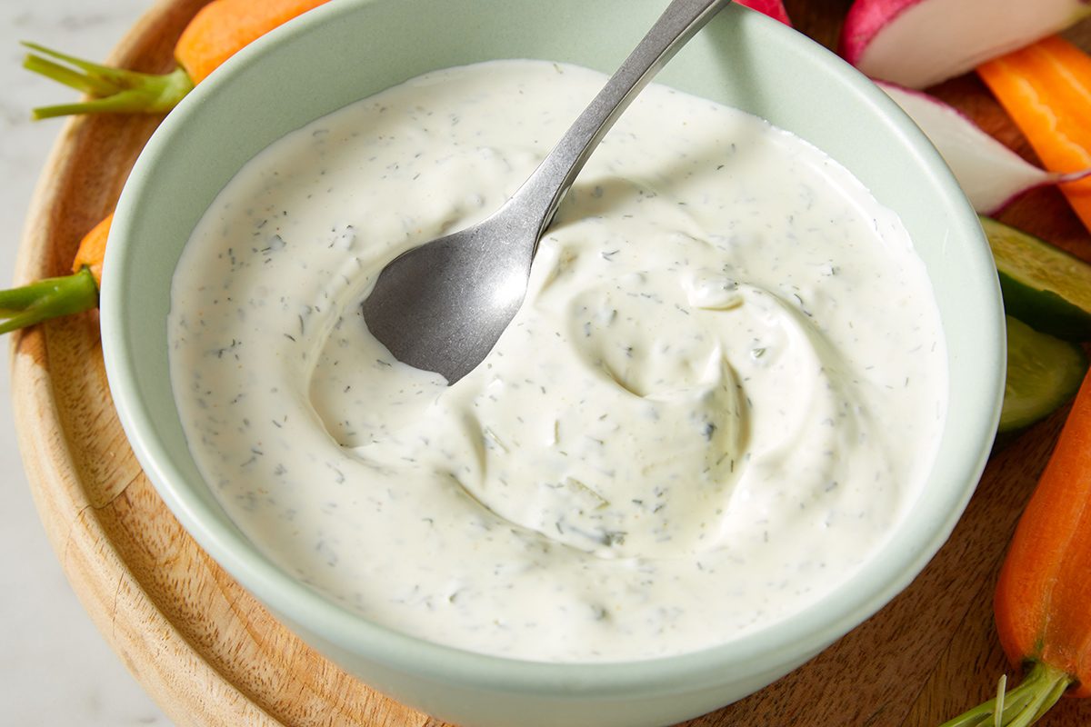 A bowl of creamy ranch dip with herbs, with a spoon resting inside, surrounded by fresh cut vegetables.