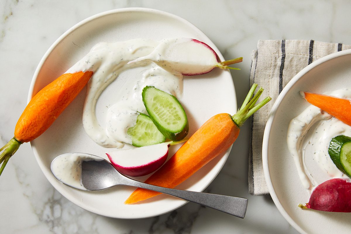 A white plate with fresh carrot, radish halves, and cucumber slices served with a swirl of creamy dip. A spoon rests on the plate, and part of a striped napkin is visible on the marble surface.