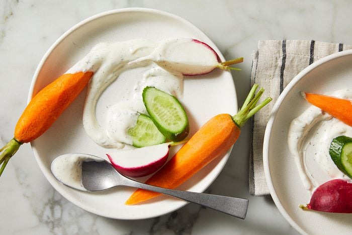 A white plate with fresh carrot, radish halves, and cucumber slices served with a swirl of creamy dip. A spoon rests on the plate, and part of a striped napkin is visible on the marble surface.