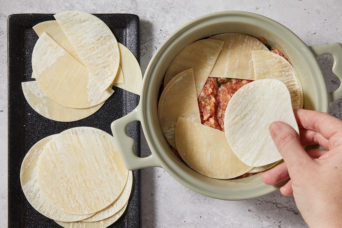 Overhead view of wonton wrappers being layered into a small baking dish over the seasoned ground pork mixture. The wrappers form the pasta-like layers of the dumpling lasagna.