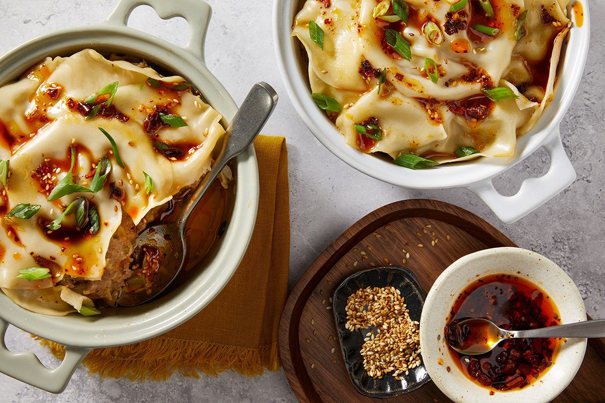 Overhead composition of dumpling lasagna served in small casserole dishes with chili crisp sauce and sesame seeds on the side. The layered wonton wrapper dish is shown ready for serving.