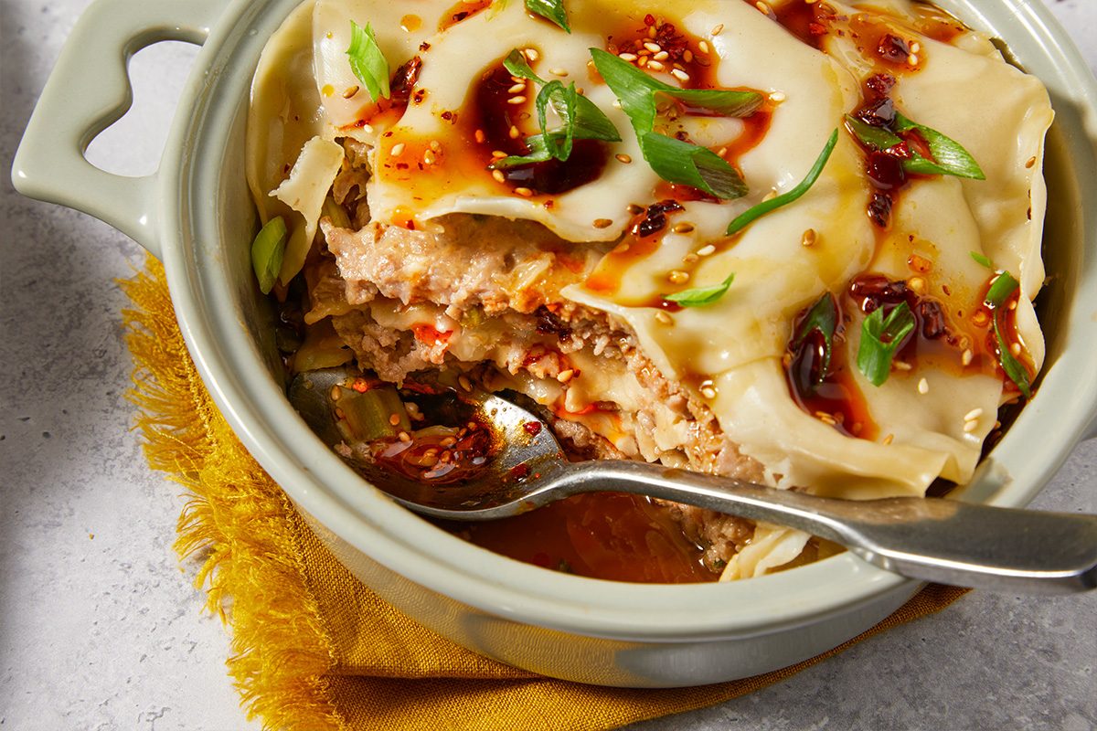 Close-up overhead view of a spoon scooping into dumpling lasagna, revealing the layered ground pork filling and soft wonton wrapper layers topped with chili crisp and green onions.