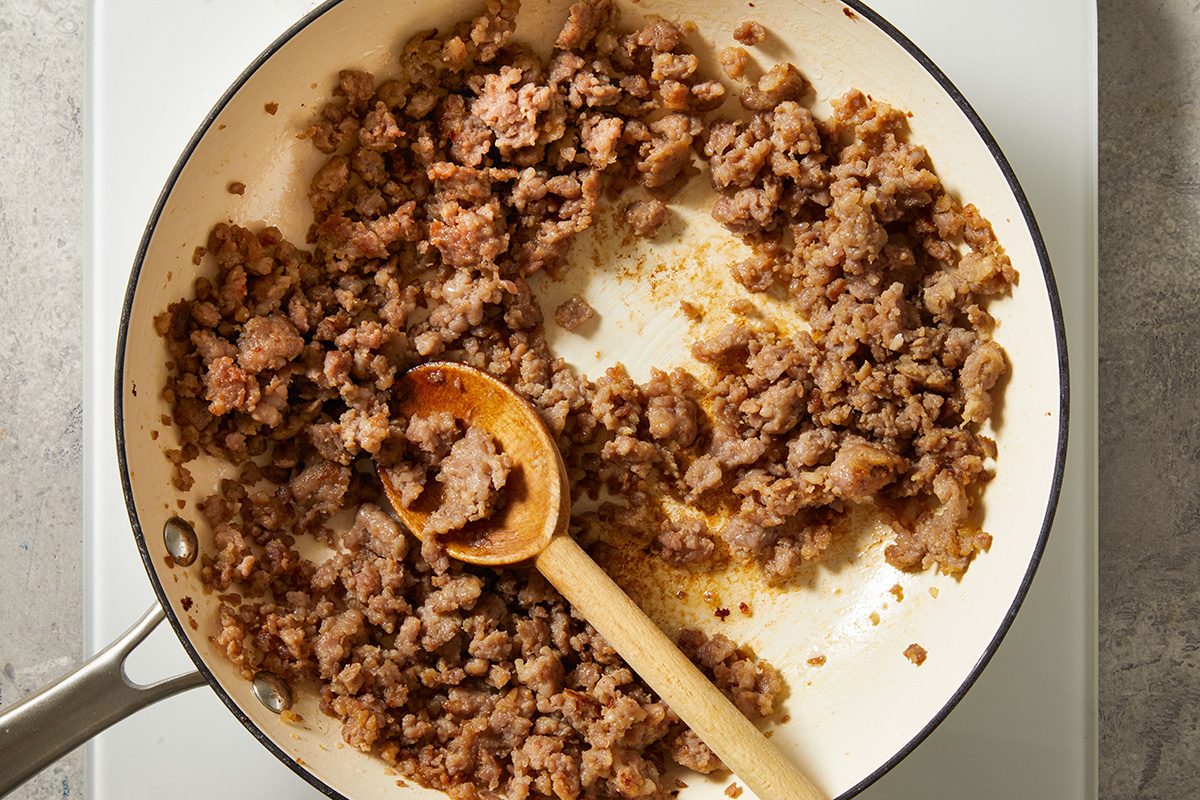 Cooked ground beef in a white skillet with a wooden spoon resting on top, sitting on a stovetop.