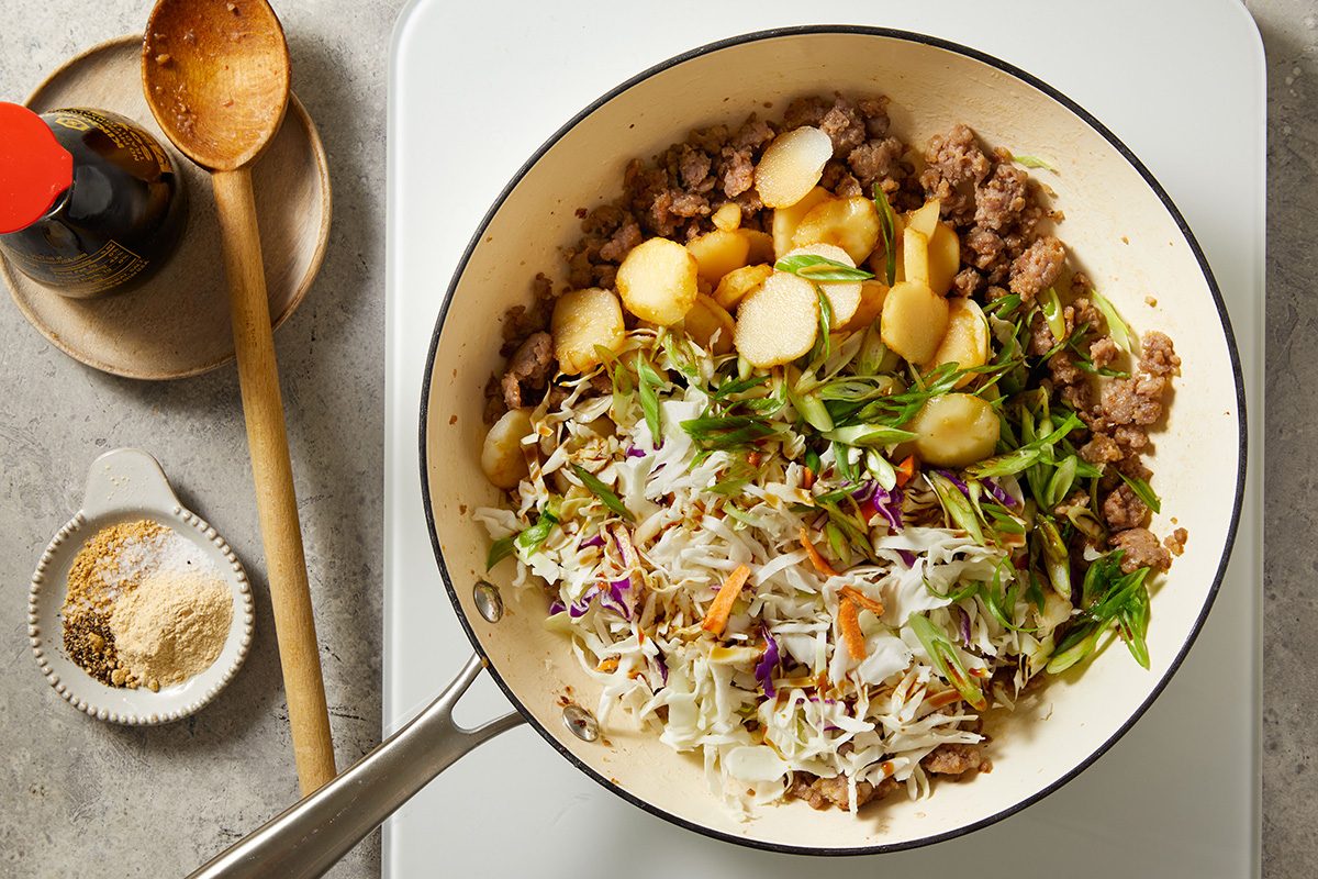 A white skillet on a stove contains cooked ground meat, sliced water chestnuts, chopped green onions, and shredded vegetables. Next to the pan are seasonings, a wooden spoon, and a bottle of soy sauce.