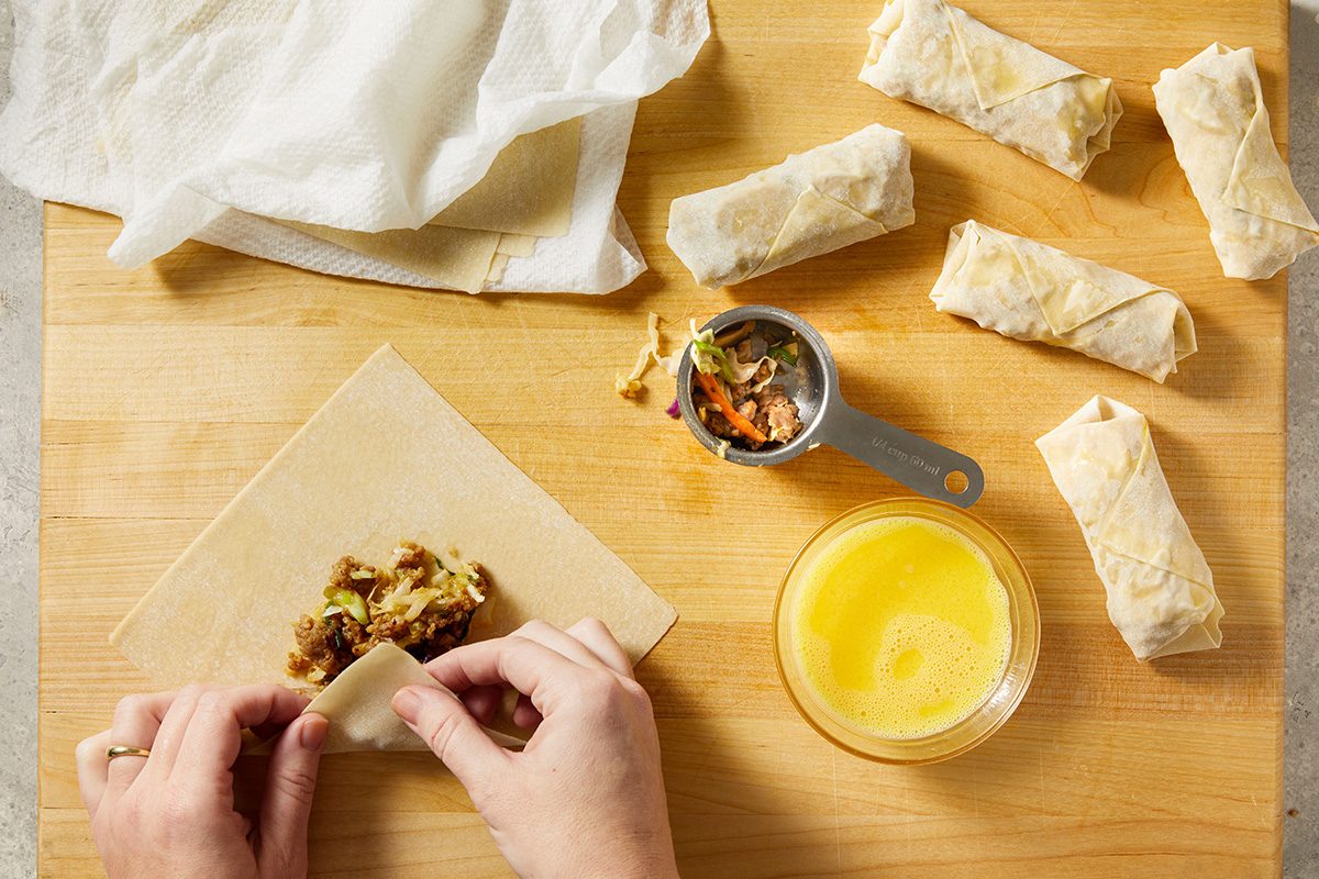 Hands rolling a spring roll wrapper with filling on a wooden surface. Nearby are finished rolls, a bowl of egg wash, a measuring cup with filling, and a cloth covering more wrappers.