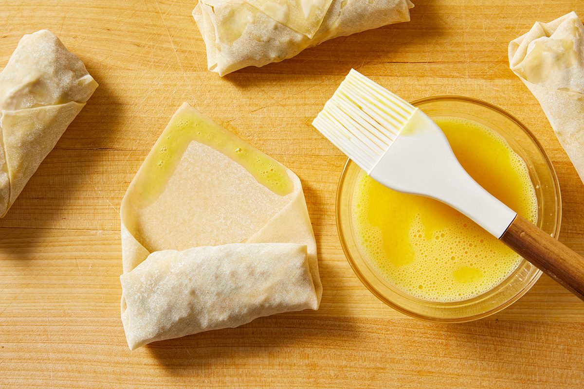 A pastry brush rests in a bowl of egg wash next to partially folded spring rolls on a wooden surface, with one spring roll being brushed with the egg wash.