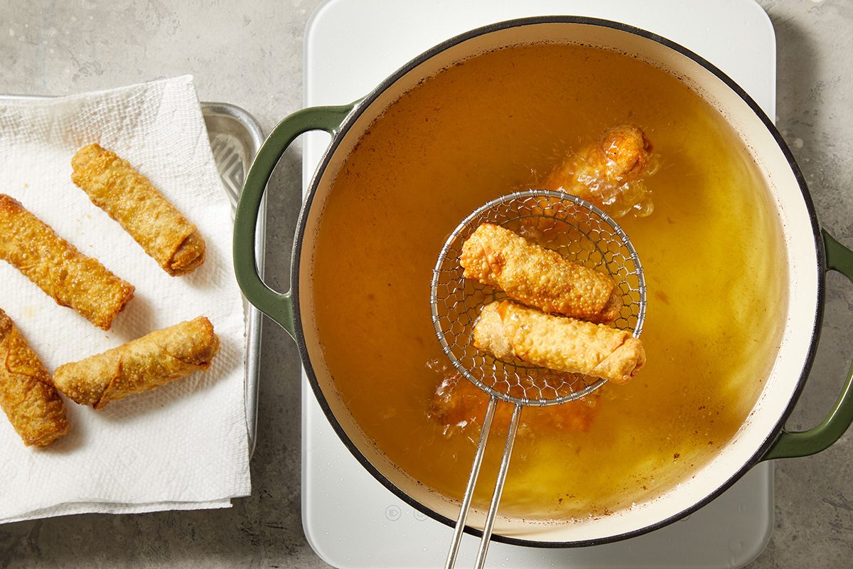 A wire skimmer lifts golden-brown egg rolls from hot oil in a pot, while cooked egg rolls rest on a paper towel-lined tray beside the pot.