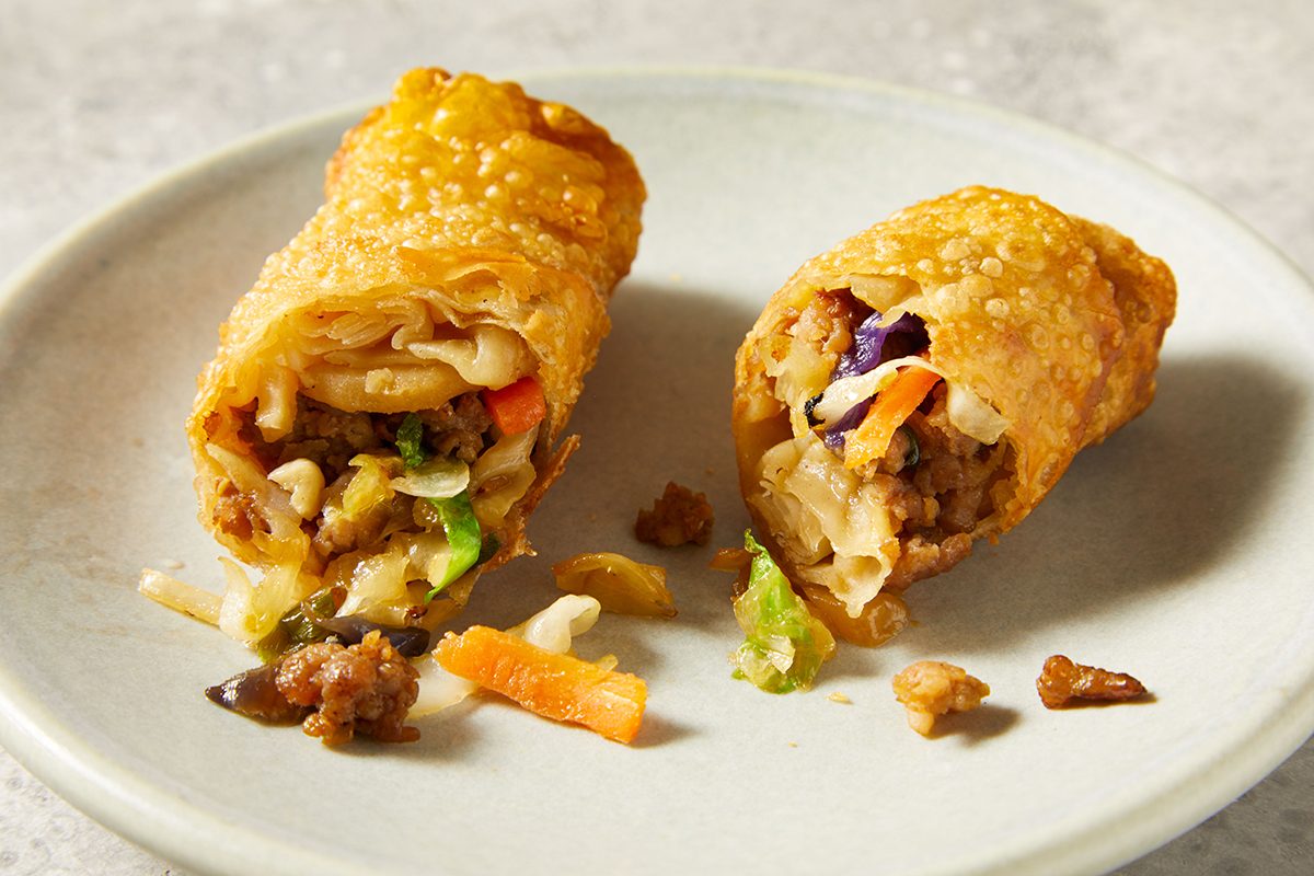 A golden, crispy egg roll cut in half on a plate, showing a filling of vegetables, ground meat, and noodles spilling slightly onto the plate.