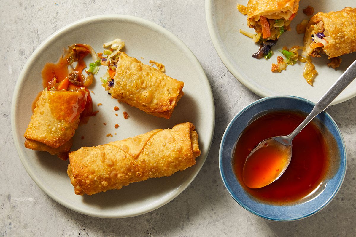 Two plates with golden-brown egg rolls, some cut open to reveal vegetable filling, are next to a small bowl of red dipping sauce with a spoon on a light-colored surface.
