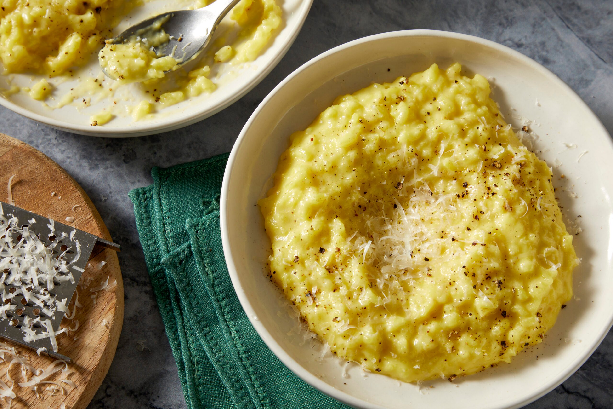 Overhead push and pulls image of a finished bowl of saffron risotto topped with grated Parmesan cheese and served with risotto cooking tools nearby.