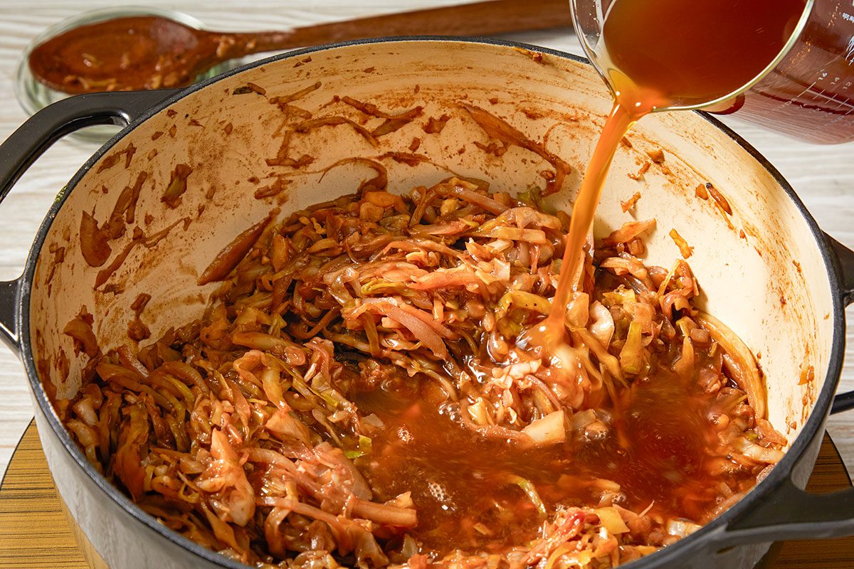 Overhead horizontal image of beef stock being poured into the cabbage and onion mixture in a Dutch oven.
