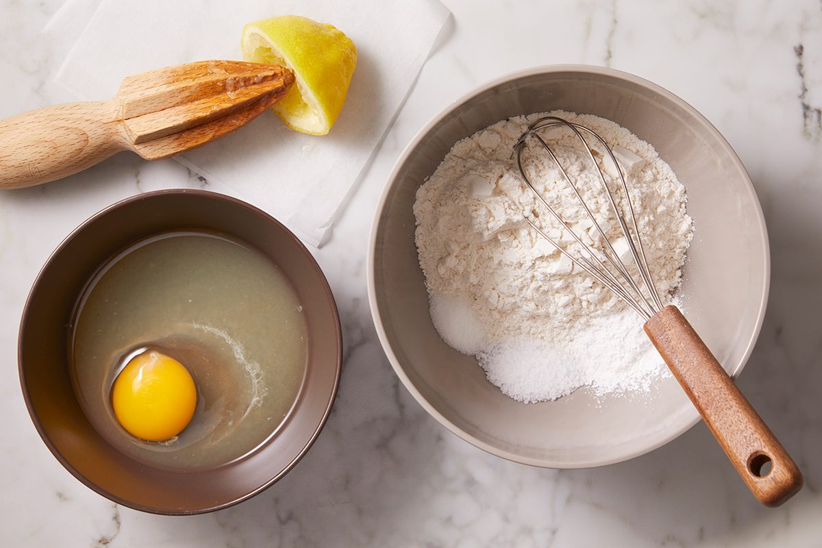 A bowl with flour and a whisk, a bowl with an egg and liquid, and a lemon half with a wooden juicer on a marble surface.