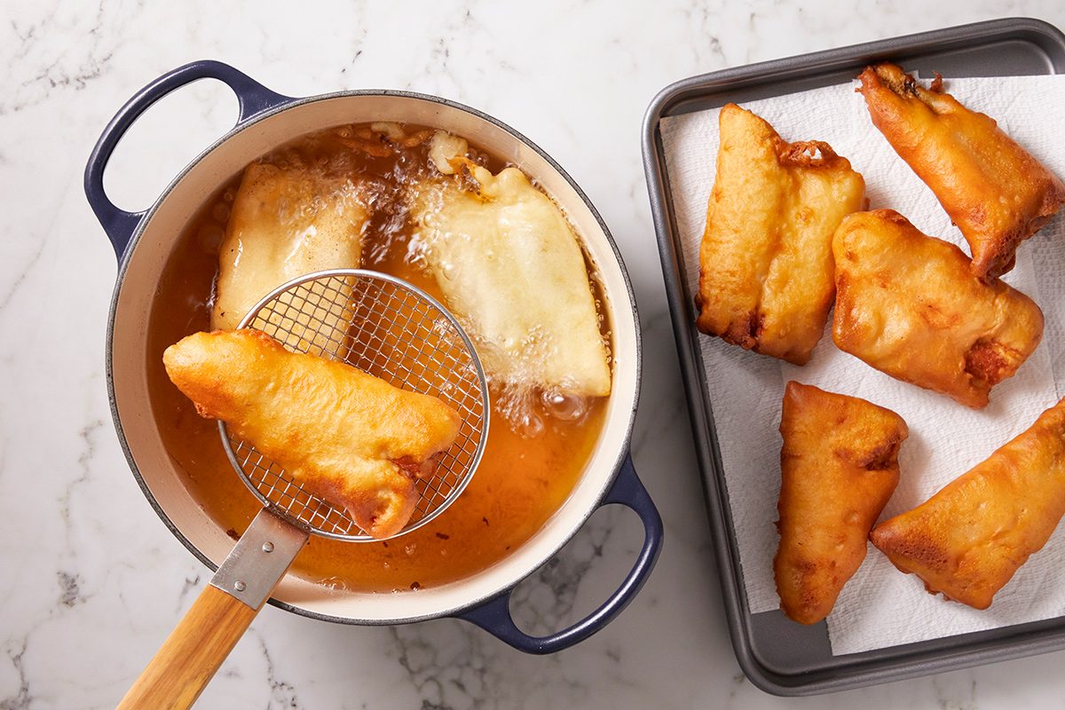 A pot of hot oil with battered fish being fried; one piece is being lifted out with a slotted spoon. Cooked golden fish pieces rest on a tray lined with paper towels.