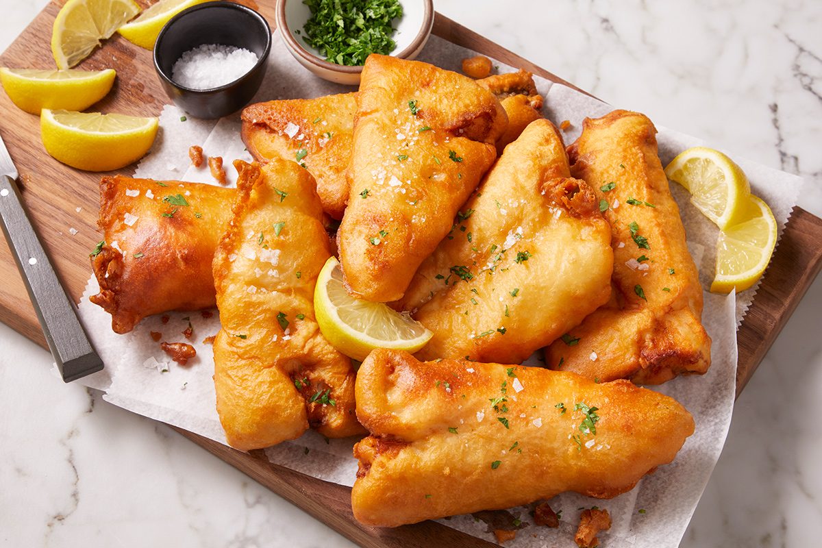 Golden, crispy battered fish fillets are arranged on a wooden board with lemon wedges, a bowl of salt, a bowl of chopped herbs, and a knife on the side, all on a sheet of parchment paper.