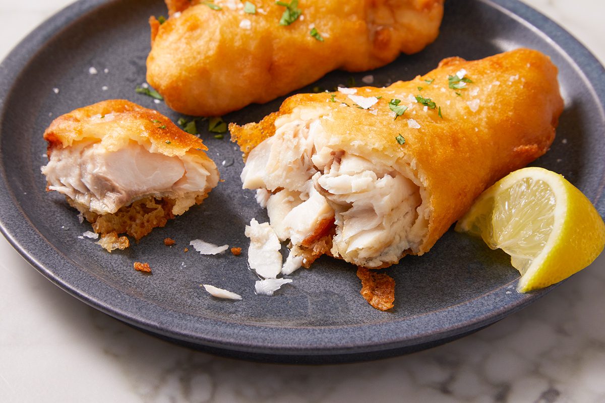 A plate with crispy battered fish fillets, one of which is cut open to show the flaky white fish inside, garnished with herbs and served with a lemon wedge.