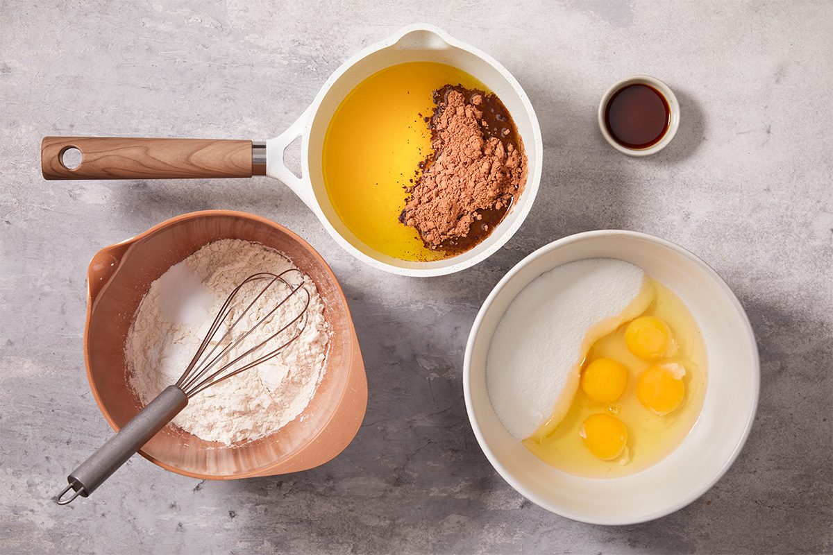 Overhead horizontal image of ingredients arranged for Frosted Fudge Brownies including cubed butter, baking cocoa, eggs, flour, and vanilla extract in individual bowls on a light surface. This step-by-step preparation image highlights the measured ingredients before mixing for a rich, fudgy brownie base.