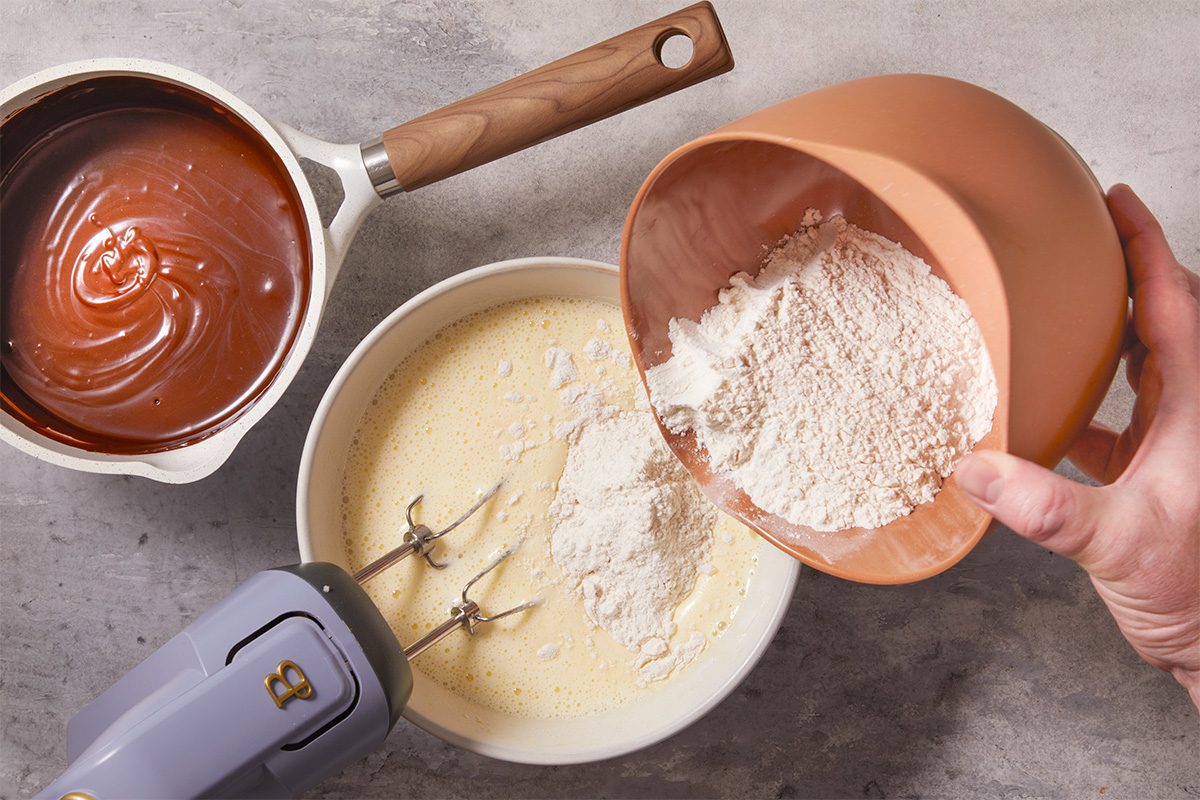Overhead horizontal step-by-step image of brownie batter being mixed, showing flour added to a cocoa-butter mixture with a hand mixer in progress. This image captures the process of combining wet and dry ingredients for Frosted Fudge Brownies.