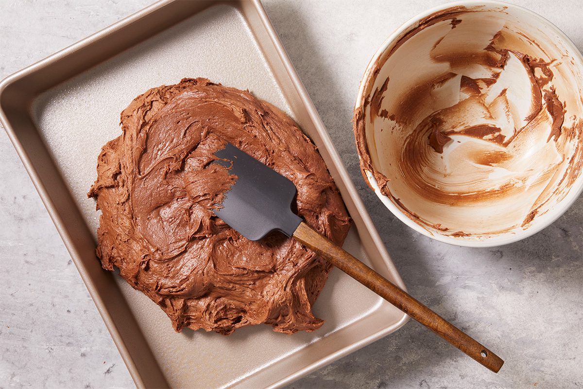 Overhead horizontal image of thick chocolate brownie batter being spread evenly into a parchment-lined baking pan with a spatula. This step-by-step image highlights preparing the batter for baking in the Frosted Fudge Brownies recipe.