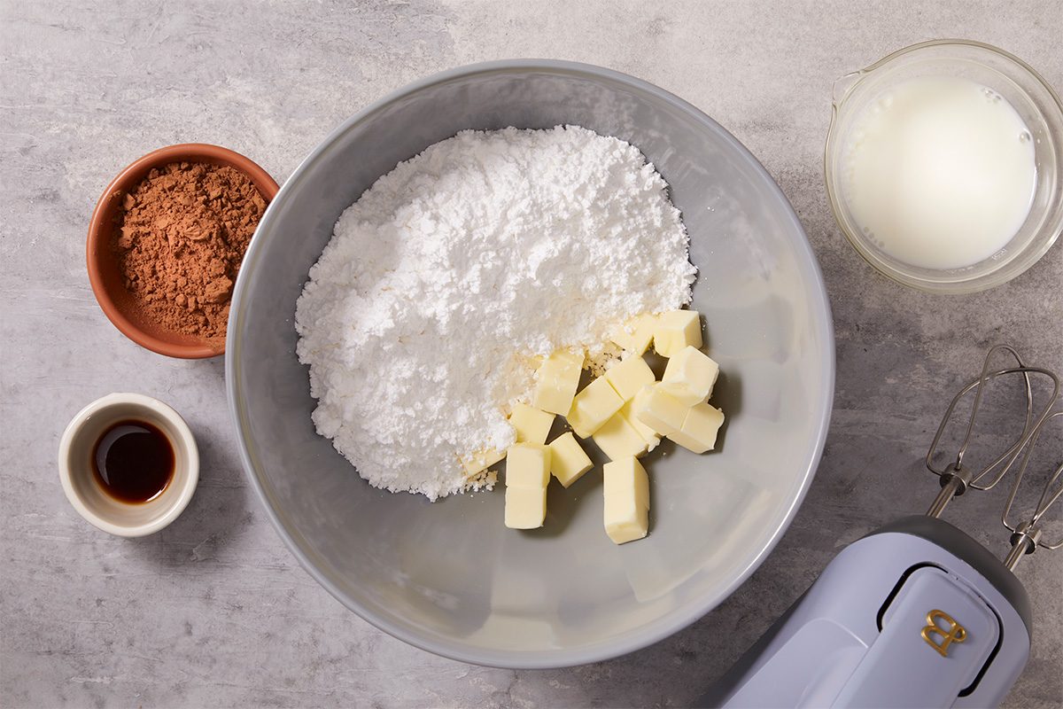 Overhead horizontal image of frosting ingredients including butter, confectioners’ sugar, baking cocoa, vanilla extract, and milk in bowls ready to be mixed. This step-by-step image shows preparation for the chocolate frosting used in Frosted Fudge Brownies.