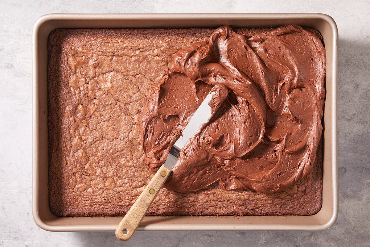 Overhead horizontal image of freshly baked brownies in a pan being topped with thick chocolate frosting spread smoothly across the surface. This push and pulls image highlights the finishing stage of Frosted Fudge Brownies before slicing.