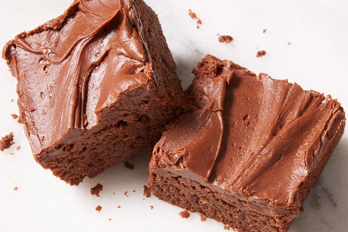 Overhead horizontal push and pulls close-up image of two large Frosted Fudge Brownie squares highlighting the thick fudgy interior and smooth chocolate frosting layer. Crumbs and texture detail emphasize the richness of this homemade dessert.