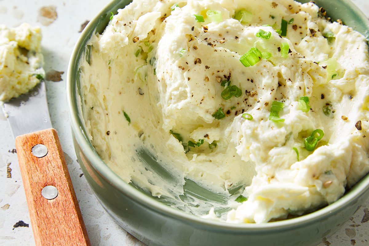 A bowl of creamy whipped cream cheese mixed with chopped green onions and cracked black pepper, with a spreading knife resting nearby.