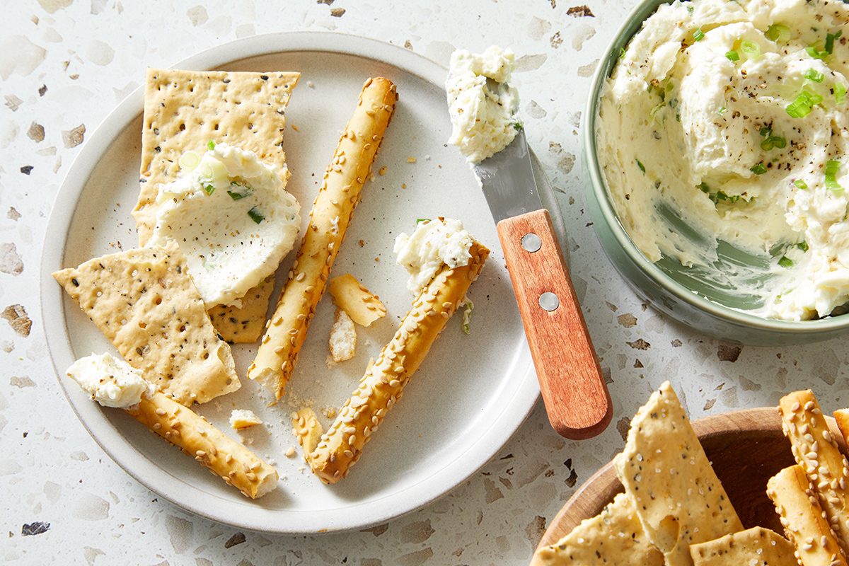 A plate with broken crackers and breadsticks spread with creamy cheese sits next to a bowl of cheese and a knife with a wooden handle on a speckled countertop.