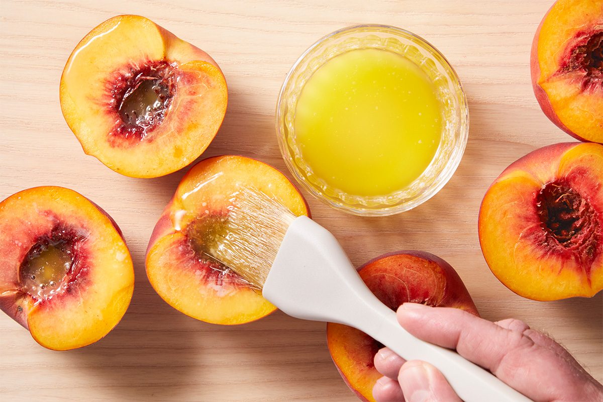 Overhead shot of halved peaches being brushed with melted butter using a pastry brush. A bowl of butter sits nearby, with the peach pits removed and facing upward.
