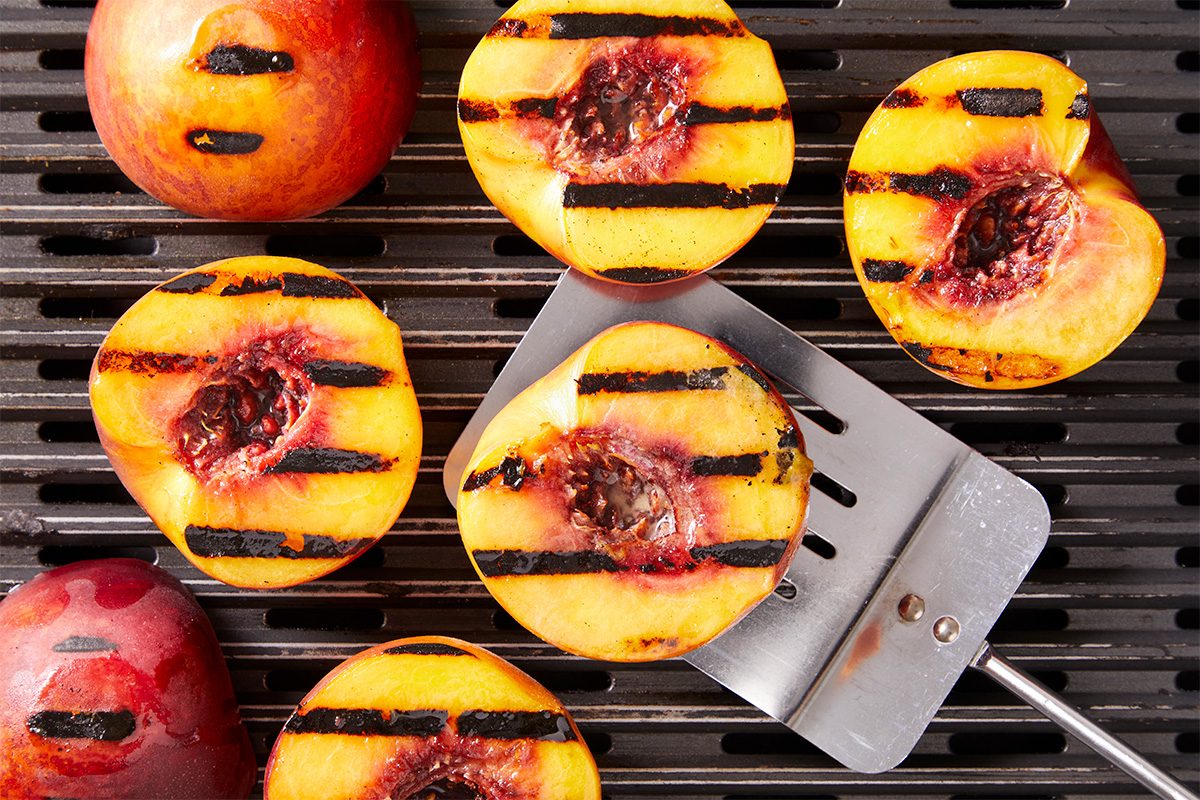 Overhead shot of halved peaches cooking on a grill, showing defined char marks. A metal spatula lifts one peach half from the grate.