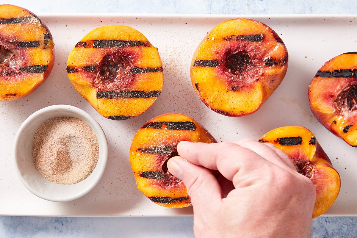 Overhead shot of grilled peach halves arranged on a tray while a hand sprinkles a cinnamon-sugar mixture over the warm fruit. A small bowl of the mixture sits nearby.