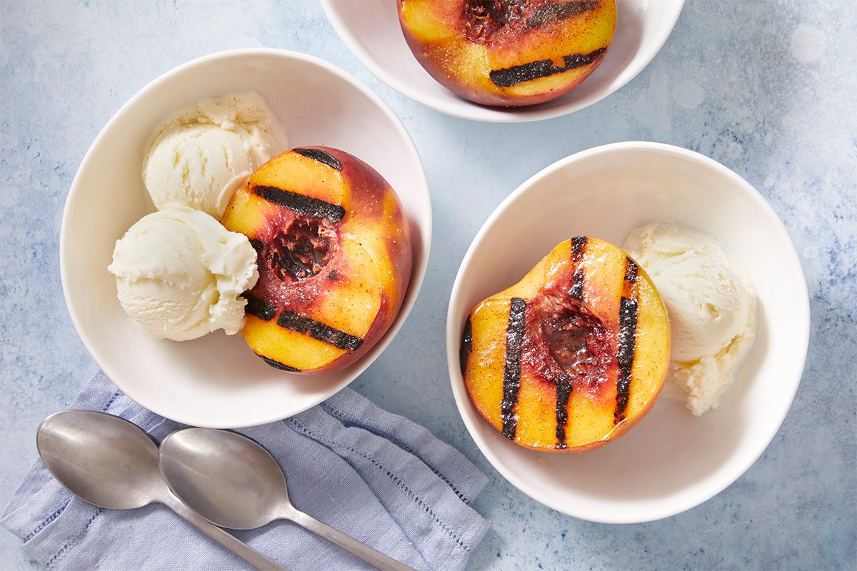 Overhead shot of two bowls of Grilled Peaches served with scoops of vanilla ice cream. Three spoons rest beside the bowls on a white surface.