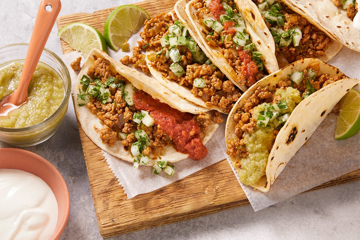 Overhead shot of Ground Pork Tacos stuffed with spiced ground pork, fresh cilantro, and crumbled toppings, served with lime wedges and sauce on a wooden surface.