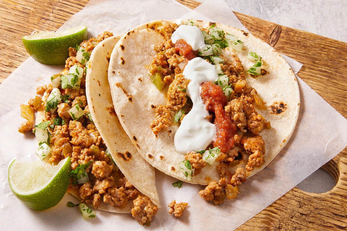 Close-up shot of Ground Pork Tacos showing browned pork crumbles topped with creamy sauce and herbs, folded in soft tortillas with lime wedges alongside.