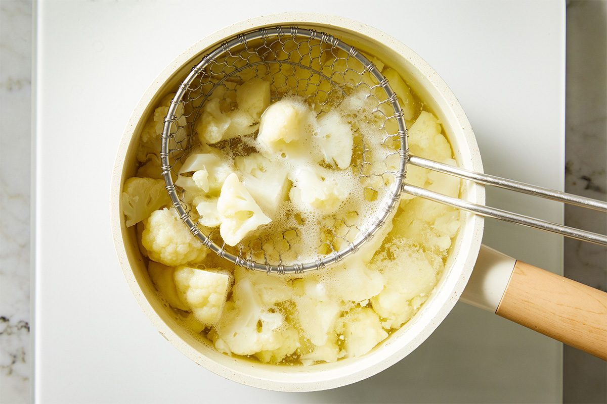 Overhead shot of steamed cauliflower florets in a saucepan, being lightly mashed with a fork, creating a chunky base for the casserole.