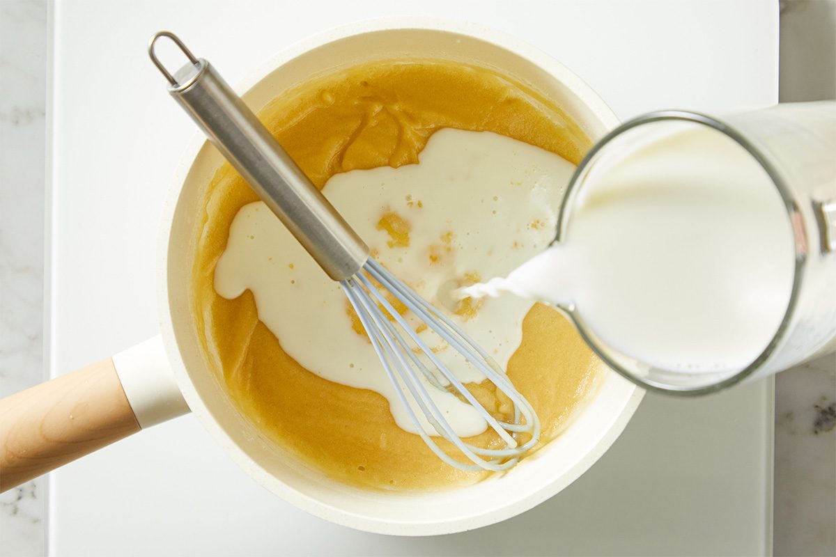 Overhead shot of milk being poured into a bowl of smooth, yellow cheese sauce while a whisk rests inside, showing the sauce being loosened.