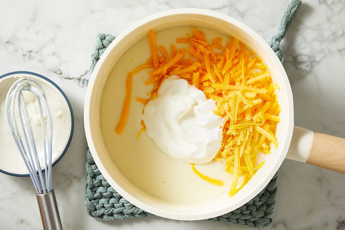 Overhead shot of a mixing bowl containing creamy sauce topped with shredded cheddar cheese and a dollop of sour cream, with a whisk nearby.