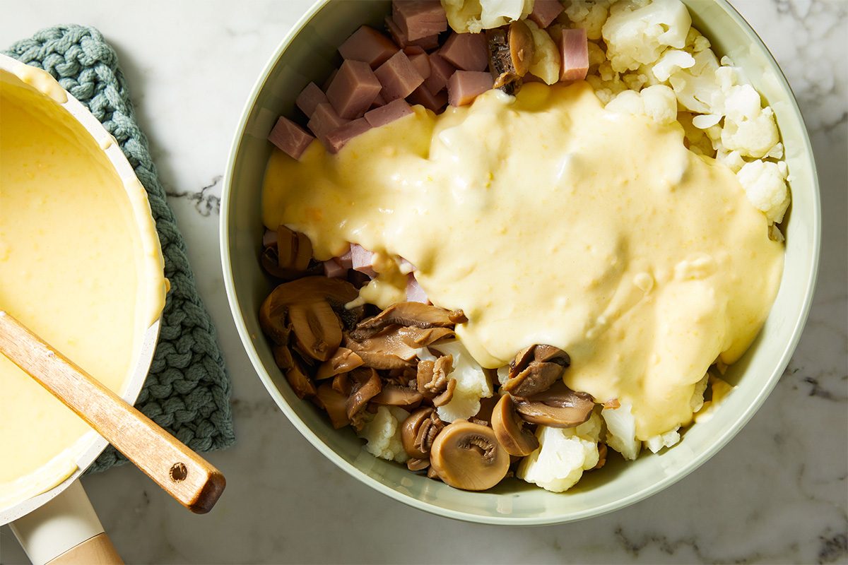 Overhead shot of a bowl filled with cauliflower mixture, diced ham, and creamy sauce, partially stirred together.