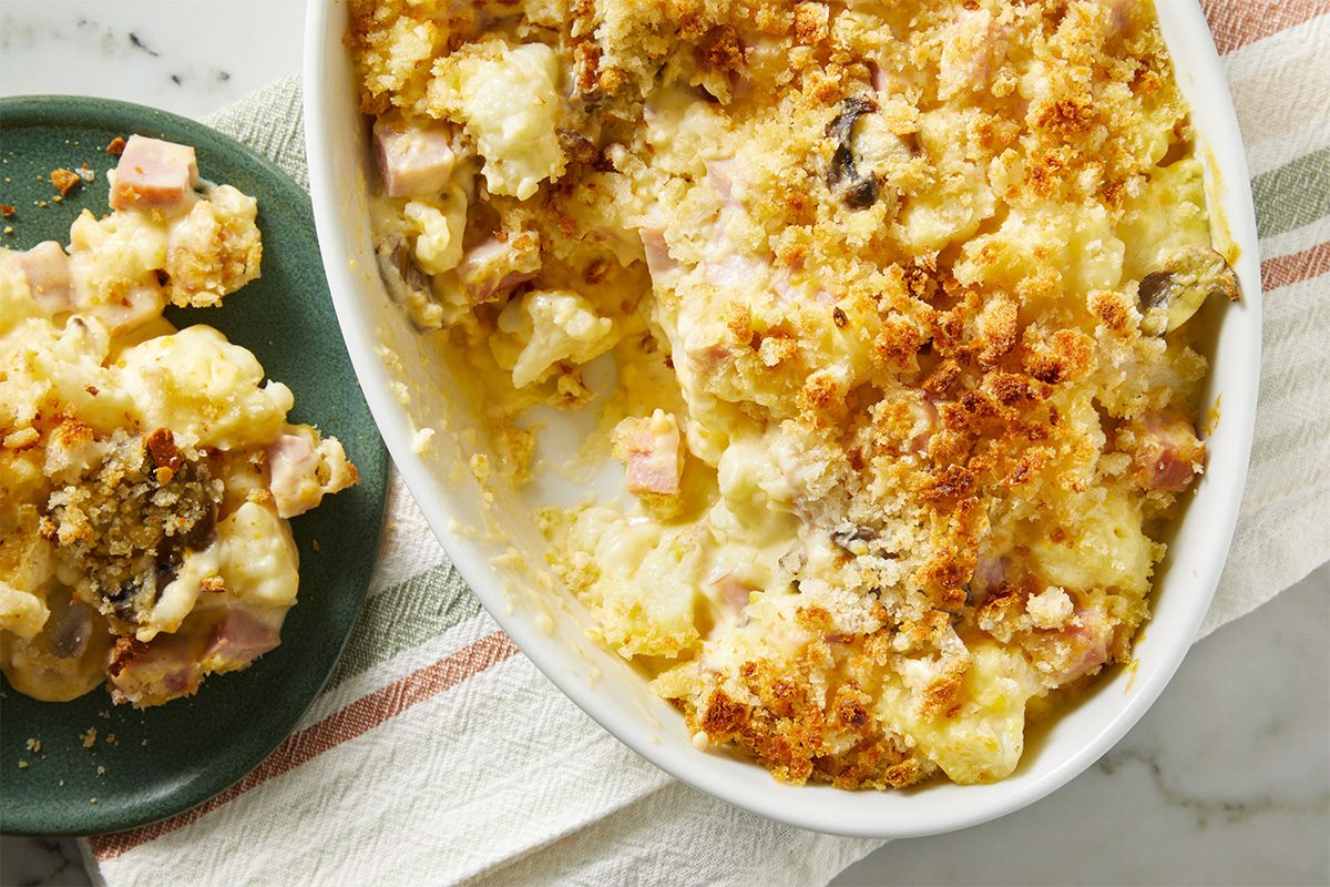 Overhead shot of a Cauliflower Ham Casserole, serving spoon scooping a portion of the baked casserole from the dish, revealing chunks of ham and cauliflower.
