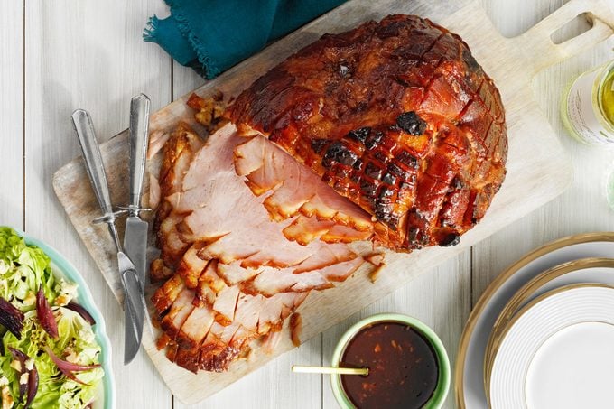 overhead shot of Spice Plum Baked Ham with knives next to salad bowl, plates and small bowl of sauce
