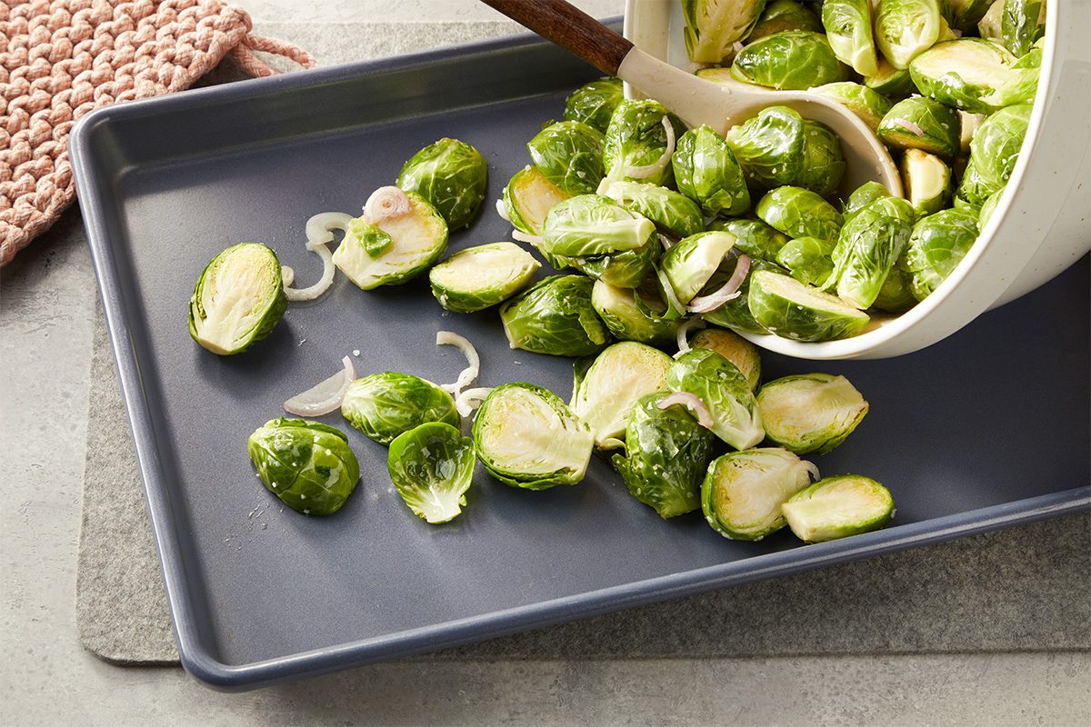 Overhead horizontal image of halved Brussels sprouts and sliced shallots being spread onto a sheet pan for roasting.
