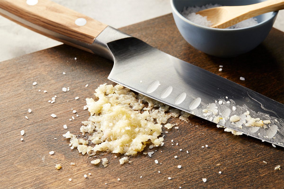Overhead horizontal close-up of minced garlic on a cutting board next to a chef’s knife.