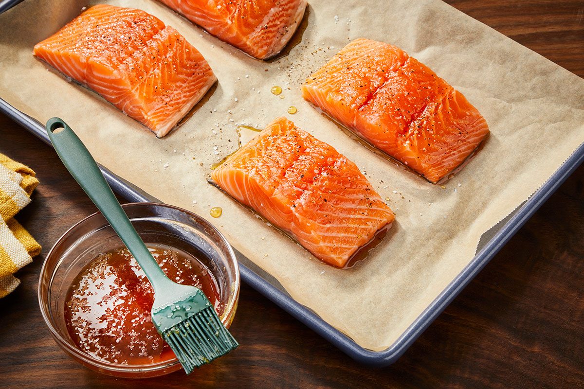Overhead horizontal image of salmon fillets arranged on a sheet pan with seasoning and glaze nearby