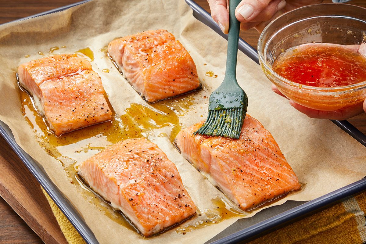Overhead horizontal image of salmon being brushed with hot honey glaze on a sheet pan