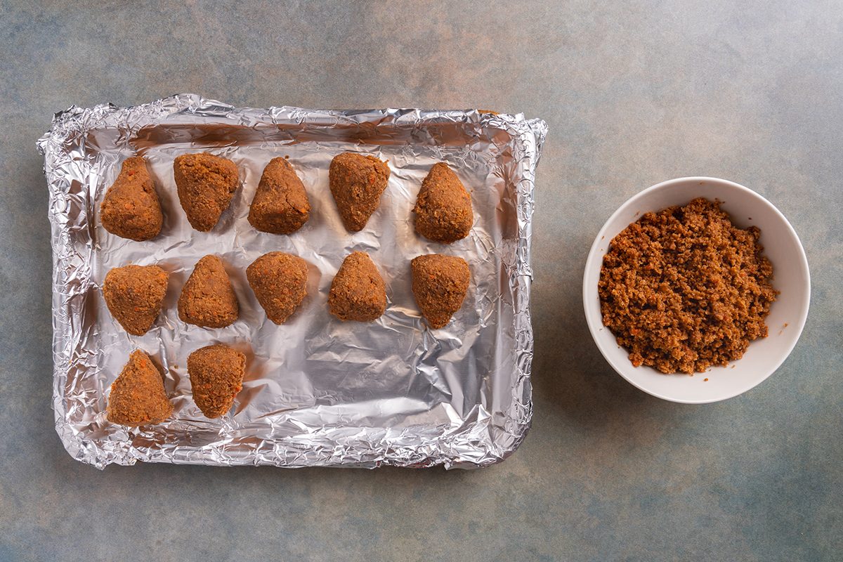 A foil-lined baking tray with twelve breaded, cone-shaped snacks is next to a white bowl filled with brown, crumb-like coating, all on a gray countertop.
