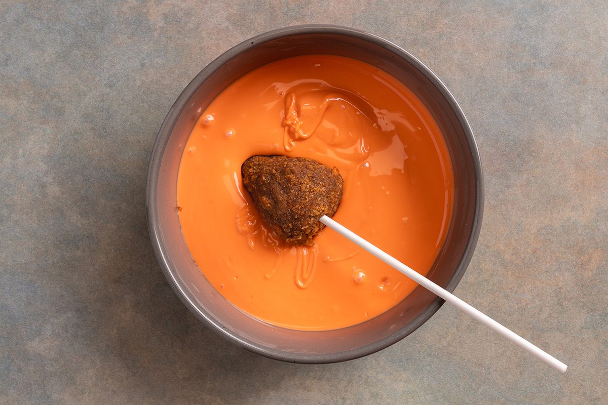 A chocolate cake pop on a stick being dipped into a bowl of orange melted candy coating, with some coating dripping off, on a gray surface.