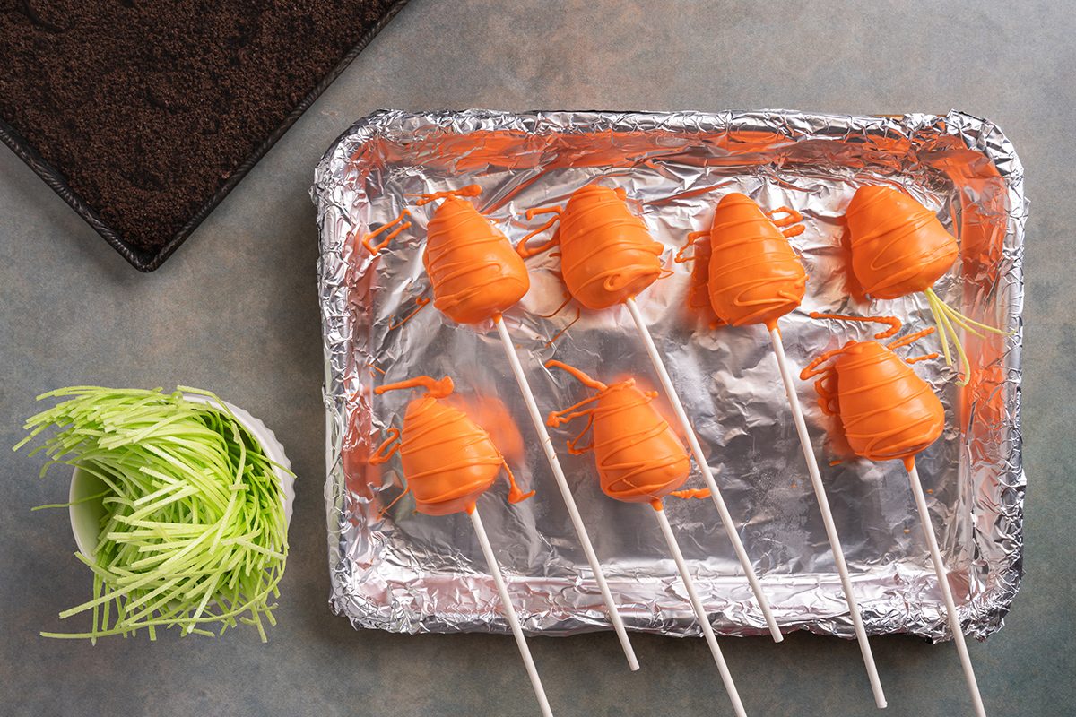 Seven cake pops shaped like carrots, covered in orange coating, are arranged on a foil-lined baking tray. A bowl of green shredded candy and a black container are nearby on a gray surface.