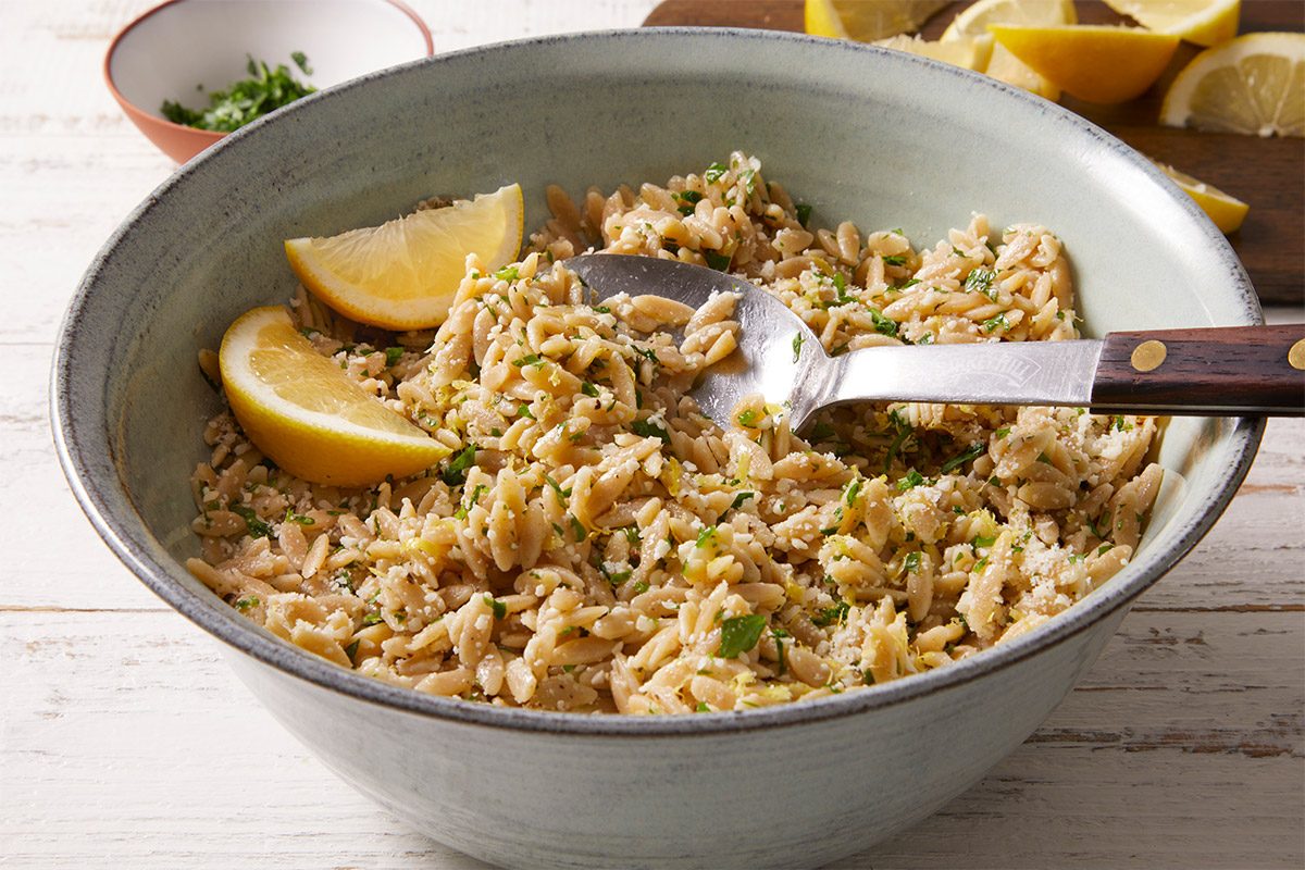 3/4 shot of a bowl of cooked lemon parmesan orzo, garnished with chopped herbs and lemon wedges, with serving spoons.