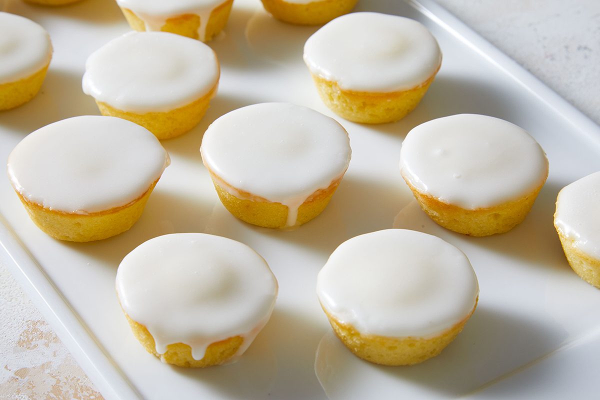 Twelve small, round yellow cakes topped with smooth white icing are arranged neatly on a white rectangular tray.