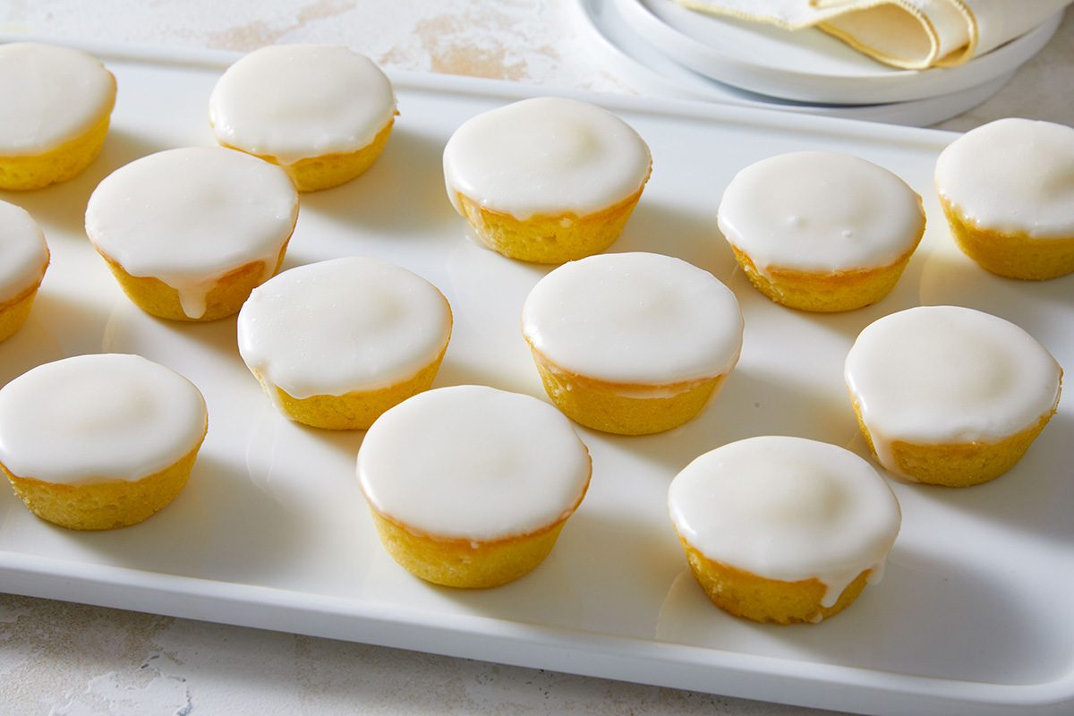 A white tray holds fifteen small round cakes, each topped with a smooth layer of white icing. Plates and napkins are visible in the background.