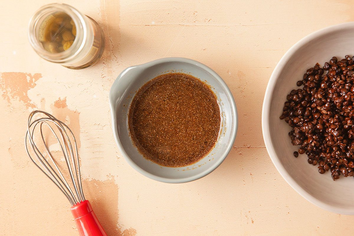 Overhead horizontal image of vinaigrette dressing with whisk alongside cooked black lentils on a neutral background.